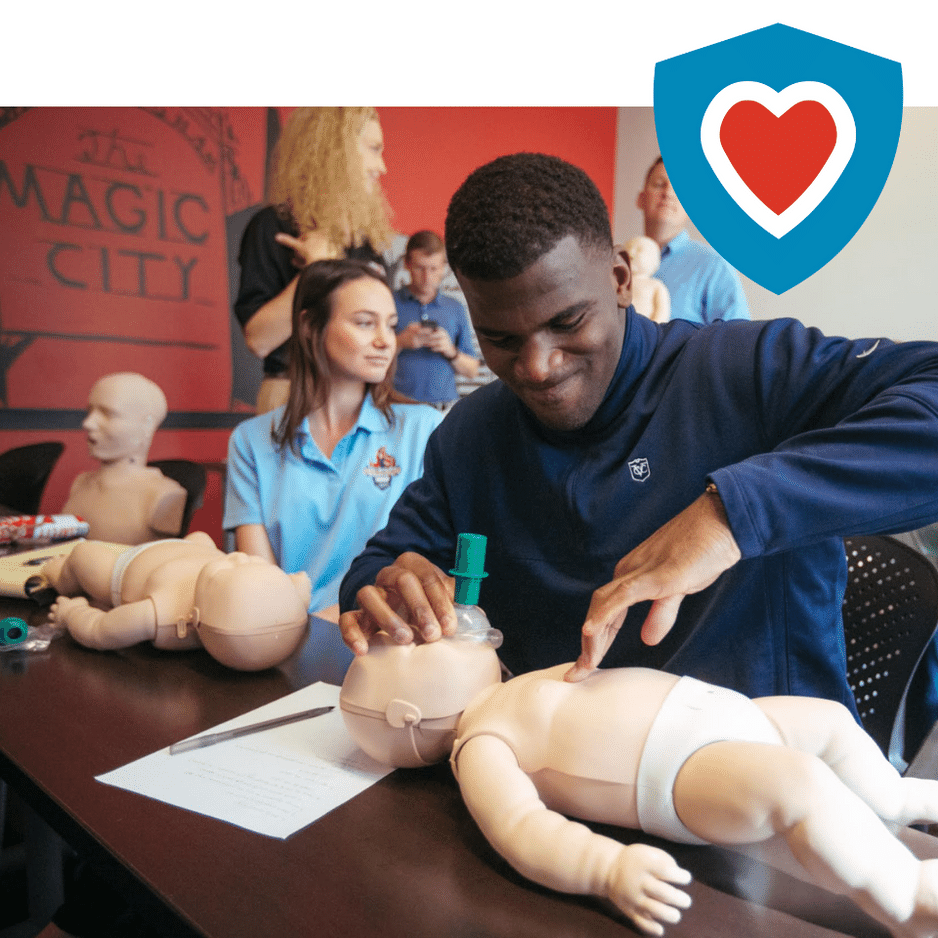 A man is diligently performing CPR on an infant CPR manikin placed on a table in front of him. His focus and care are evident as he demonstrates the life-saving technique. To his side, a young girl is sitting, her smile radiating interest and encouragement. Behind them, a group of attentive onlookers stands, observing the demonstration closely.