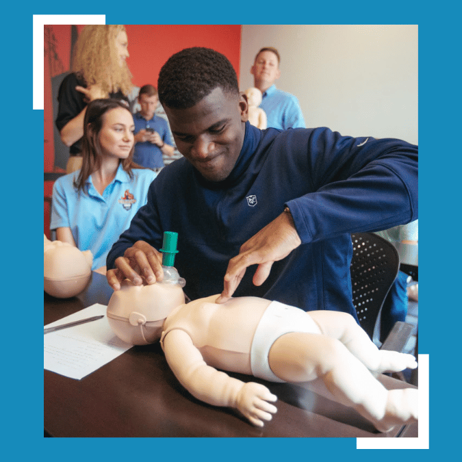 A man is diligently performing CPR on an infant CPR manikin placed on a table in front of him. His focus and care are evident as he demonstrates the life-saving technique. To his side, a young girl is sitting, her smile radiating interest and encouragement. Behind them, a group of attentive onlookers stands, observing the demonstration closely.